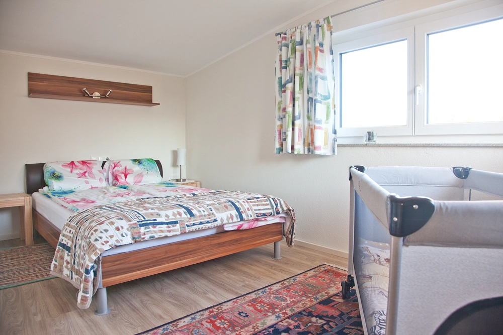 Ferienwohnung Greiler – Interioraufnahme; Schlafzimmer in weiß mit Holz, heller und freundlicher Raum mit Kinderbett, strukturierte Raumaufteilung.